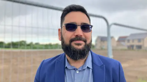 JOANNA TAYLOR/BBC Atteq Nasir smiling at the camera. He is wearing dark sunglasses, a navy blazer and a blue shirt. Behind him is a patch of mud - where the building will be erected - surrounded by a metal fence. 