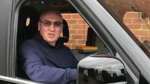 Mark White A bald man wearing a black zip-up jumper and black sunglasses sits in the driver's seat of a black cab.