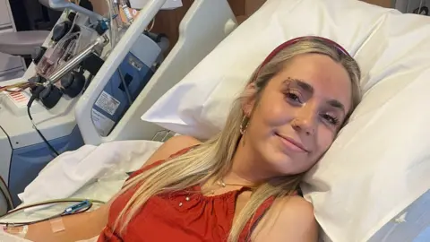 Beth Bowyer Beth Bowyer is laying down in a hospital bed and is wearing a red top and headband. She's smiling at the camera.