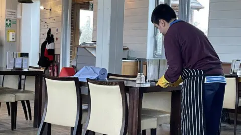 SWRAC Tommy, a young man with black hair, is wearing a maroon coloured jumper and blue sweatpants with a striped apron and marigold gloves as he leans over a restaurant table to clear it