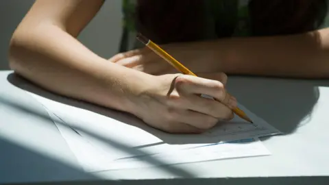 Getty Images A person holding a yellow pencil and writing on a page. The picture only shows the person's arm and hand.