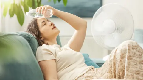 A woman reclines on a sofa next to a fan and with a bottle of water pressed to her forehead