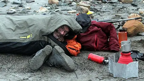 Mike Newman Prof John Marshall wild camping on rocky ground on an expedition to Spitsbergen in 2023. He smiles from a gap in his sleeping bag, which mostly covers his head. A pair of mud-encrusted boots, a small gas stove and other equipment are beside him.