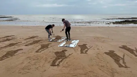 Phil Harrison/BBC Silhouettes of soldiers being installed onto Stone Bay beach 