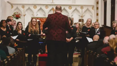 A-Teck Events A group of women singers holding folders, wearing red poppies and singing in a church and facing the congregation. One of them is in police uniform. Their musical director is standing in front of them, wearing a red and black jacket and black trousers. There are pews on either side filled with people.