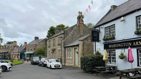 A row of stone buildings face into a village square. The nearest building is a pub, which painted blue and has a sign reading "Washington Arms" on the front. Colourful bunting is strung up from lamppost to lamppost.
