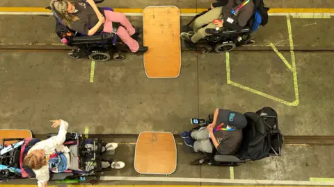 Wheelchair users are positioned as if they were sitting in the train carriage. There are four students visible from above and they are sitting next to wooden tables on the floor