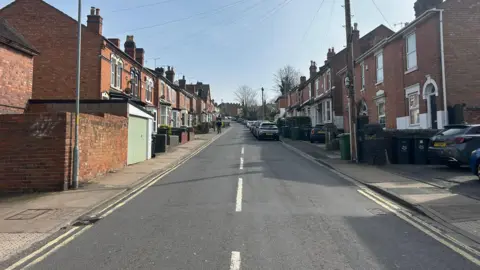 A general view of a residential street with terraced houses on either side of the road and cars parked on the right side of the road.