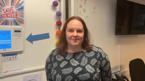 A woman wearing a grey top with a leaf pattern on it. She is standing against a white wall in what appears to be an office, above her right shoulder a poster that says Save The Hive is hanging on the wall