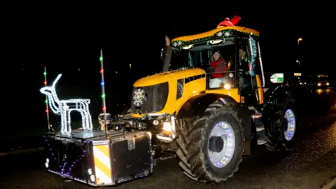 A yellow tractor in the dark decorated with festive lights, including a reindeer, and the driver is smiling to camera