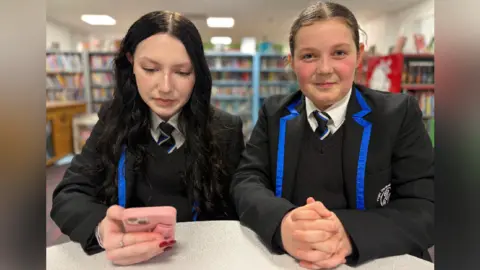 Two girls wearing school uniform with black blazers and blue edging standing together in a library with one holding a pink mobile