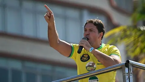 Alexandre Ramagem points with his finger with one hand while holding a microphone in the other hand as he is speaking at a demonstration in support of former Brazilian President Jair Bolsonaro at Copacabana Beach in Rio de Janeiro on 3 August, 2025. He is wearing a Brazil football shirt with several stickers on it. One bears a photo of ex-President Bolsonaro. 