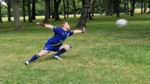 Naj Modak Mason in a blue kit with goalkeeper gloves lunges sideways across a grassy field during a training session in a park. He stretches both arms toward a fast-moving football visible in mid-air. Trees surround the open field in the background, and training markers are placed on the grass.