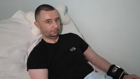 BBC A man wearing a black T-shirt and with a scar on his neck from a tracheostomy sits in a hospital bed