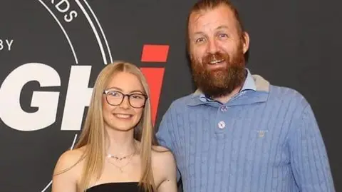 Leia Con Leia, em um vestido preto, seu pai sorriu ao lado de Stephen, que usava um saltador azul. Eles estão na frente de uma grande faixa com os 'Awards People of the Year - Armagi'