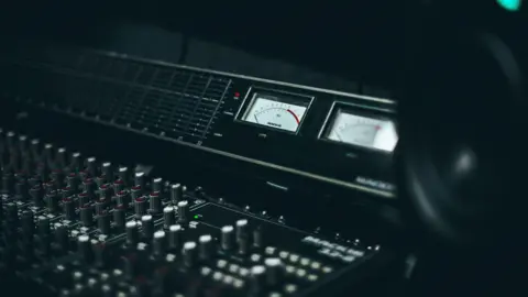 Moose Records/James Revitt Moose Records mixing desk (which used to be George Michael's) . A desk with an array of knobs and dials sits in semi-darkness.