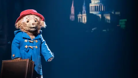 A brown bear with a blue rainjacket and red hat holds a suitcase.