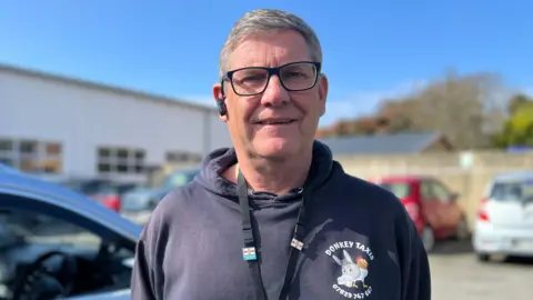 Ian Vaudin pictured with short grey hair wearing black glasses. He is wearing a black hoodie with 'Donkey Taxis' written on the front above a graphic of a donkey and a chicken. 
