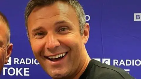 BBC A man with short grey hair and wearing a black shirt smiles while standing in front of a purple banner that says "BBC Radio Stoke".