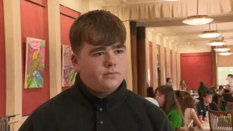 A brown haired teenager boy with thick eyebrows and a black buttoned up top speaks to the camera. 