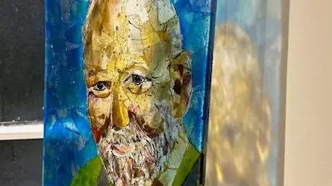 A side on view of the portrait of Graham Hall, it is made of glass, Graham has a kind smile, you can see the light of the glass reflecting on the wall in the background.