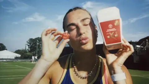 Wimbledon Aryna Sabalenka with some tennis courts in the background. She has her eyes closed ans is making a kiss shape with her mouth. In one hand she is holding up a strawberry and in the other a red carton.