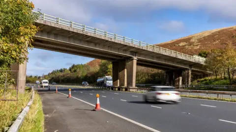 National Highways A roadside northbound view of the M6 motorway at the Lune Gorge between the Howgill Fells near Tebay, Cumbria with a number of lorries and cars travelling on the road in sunny conditions