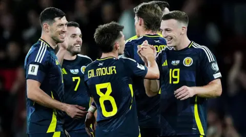 PA Scotland football players in dark blue kits with yellow accents celebrate together on the pitch, with one player wearing a jersey labeled “Gilmour” and number 8.