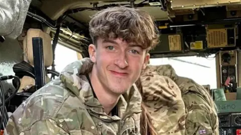 Pte Harry Johnson pictured wearing army overalls inside a plane. He has mid-length blonde hair.