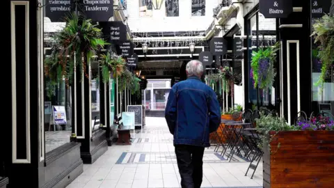 A man is pictured walking through a shopping arcade with signs and plants hanging out of the front of each retail unit