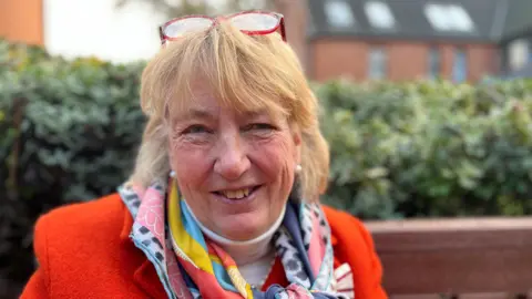 Jo Thewlis/BBC Lady Philippa Dannatt has shoulder-length, blonde hair with red glasses on her head. She is wearing a red jacket with a colourful scarf around her neck and is smiling at the camera.