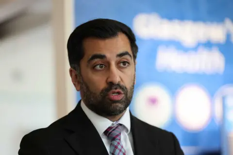 PA Media Humza Yousaf making a speech, wearing a black suit, white suit and a tartan tie. There is a blue background, which has been blurred.