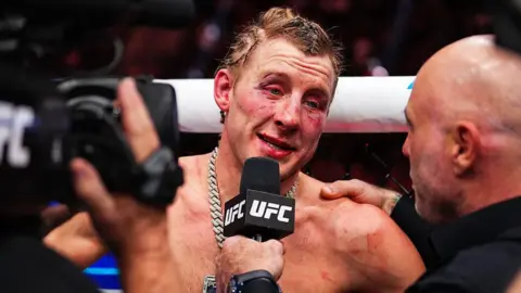 A brusied Paddy Pimblett speaks on the UFC mic. He has blood stains on his face and his eyes are red.