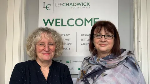 Katherine Robertson and Shelley Hunt stand in front of their white business sign which reads 'Lee Chadwick Solicitors' then below 'Welcome' in green. Katherine wears a navy blue dress with pale blue polka dots. Shelley wears a purple scarf draped across her shoulder. They smile into the camera.
