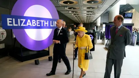 The late Queen Elizabeth II walking through the concourse of an Elizabeth line station accompanied by two men, in front of a large 'Elizabeth line' roundel sign.