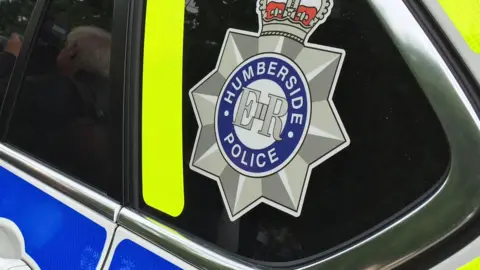 A close up of the back of a Humberside police car showing the logo with ER at the center.  The emblem is a star badge, topped with a crown, and a blue circle in the centre saying Humberside police.