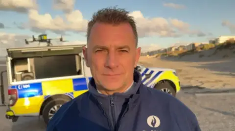 Christian De Ridder from Westkust Police in Belgium is standing in front of a police vehicle and a police drone on a beach in the coastal town of De Panne.
