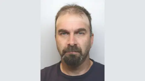 A police mugshot of Daniel Tunney. he is wearing a dark-coloured t-shirt and has thin brown hair and a beard.