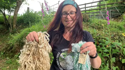 A woman with long brown hair and a woven mohair band holds up curly sheep wool that is undyed and compared with her own dyed fibre. 