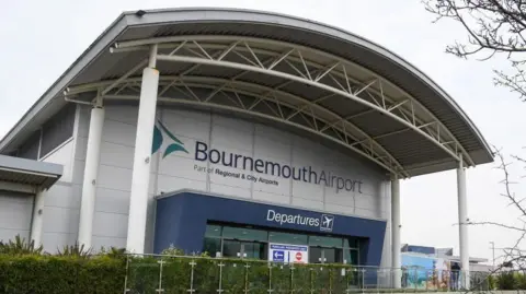 Bournemouth Airport Departure building - a grey building with a large portico held up by a number of columns and a blue surrounding to the glass entrance and a green hedge at the front.