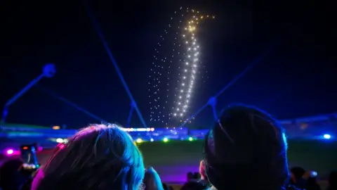 Drones make the shape of a penguin in the sky. Spectators are in the foreground looking up at the display.