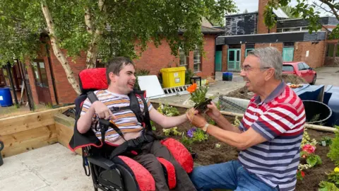 Wheelchair user Mark has short dark hair and is wearing a white and orange striped t-shirt and brown trousers. Peter has short grey hair and is wearing silver framed glasses. He is wearing a red, blue and white striped t-shirt and blue jeans. Both are next to a flower bed and are both holding a plant ready to plant.