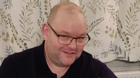A man, Gary Stanley, smiles into the camera. He is wearing black glasses and a black top. Behind him are white curtains with details of green leaves and branches.