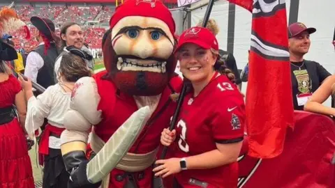 Christie Thomas Christie Thomas in a red shirt holding up a flag next to a mascot.