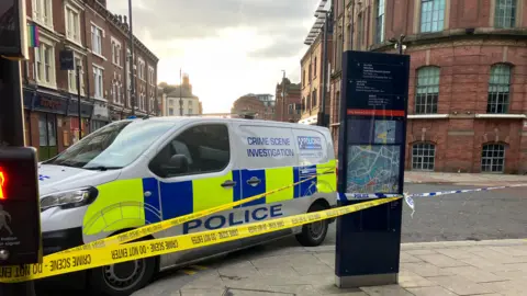 A white, yellow and blue police van parked on the corner of a street. There is yellow 'crime scene' tape around it and on the back of the van it reads 'crime scene investigation'.