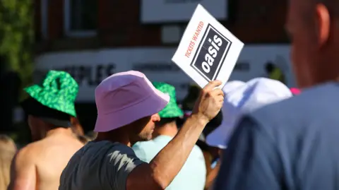 Emma Gibbs A man in a pink bucket hat holds a sign reading: Tickets Wanted Oasis. People in bucket hats walk past him