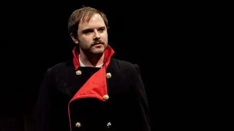 Temple Photography Peter on stage. He's wearing a black naval pea coat with strong red lapels and gold buttons.