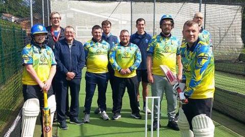 Ten men in cricket gear and blue and yellow kits standing by a wicket.