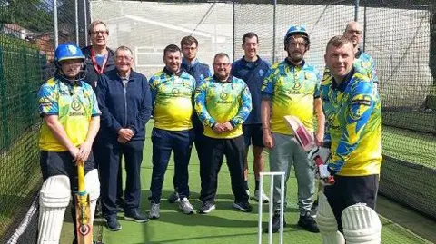 Hartlepool Borough Council Ten men in cricket gear and blue and yellow kits standing by a wicket.