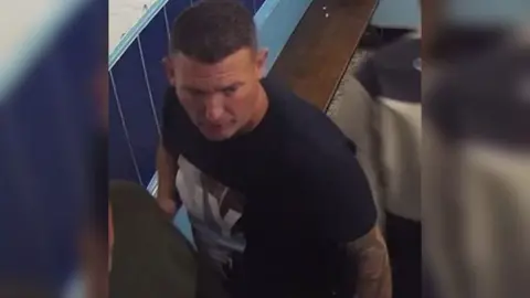 A screenshot from CCTV footage showing a man in a takeaway. He is wearing a dark coloured t-shirt with a graphic pattern on the front, has short brown hair and a tattoo sleeve on his left arm. 